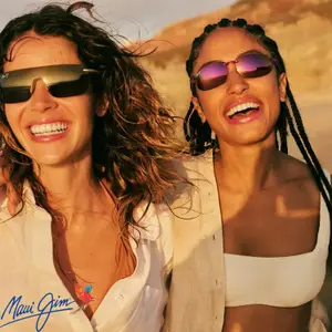 Two women in sunglasses on a beach, one with a white shirt and the other with a white crop top, both smiling and looking at the camera.