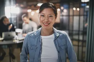 A woman in a denim jacket and glasses is smiling in an office setting.