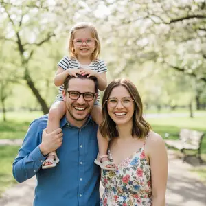 A family of three, including a little girl with glasses, are smiling and standing close together in a park with trees and grass in the background.