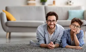 A man and a boy wearing glasses are smiling while lying on the carpeted floor in the living room.