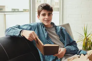 A young boy is sitting on a couch and holding a book while smiling and looking at the camera.