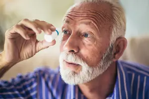 An older man with white hair and a beard is holding a bottle of eye drops near his eye.