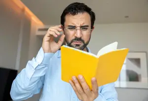 A man in a blue shirt is holding a yellow book while adjusting his glasses.