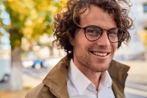 A man with curly hair wearing a white shirt and glasses, smiling at the camera on a sunny day.