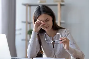 A woman sitting at a desk with a laptop and notebook, holding her forehead with one hand and her glasses with the other, looking stressed.
