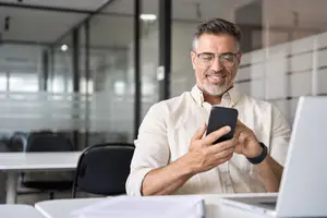 An older man wearing glasses and a white shirt is sitting in an office and looking at his phone.