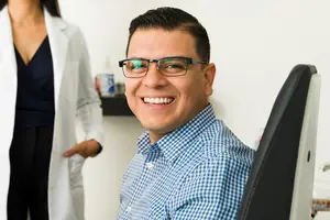 A smiling man wearing glasses and a blue checkered polo shirt sits in a chair, with a woman in a white lab coat standing behind him.