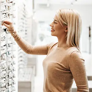 A woman wearing a brown long-sleeved shirt is smiling while looking at a wall of glasses in a store.