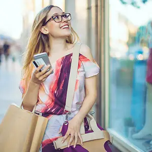 A woman with glasses and a smile is holding a phone and a bag in her hand in an outdoor setting.
