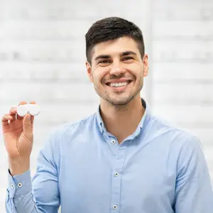 A smiling man in a blue shirt holds two white contact lenses.