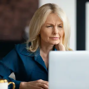 Elderly woman with blonde hair sitting in front of a computer monitor, looking sad or frustrated.
