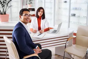 A man in a suit sits in a chair in an optometrist's office, while a woman in a white coat works at the desk in front of him.