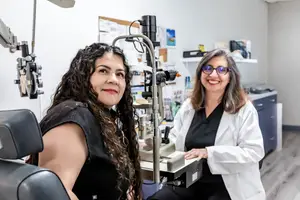 Woman with curly hair sitting in a chair in front of a woman in a white coat examining her eyes using an ophthalmoscope