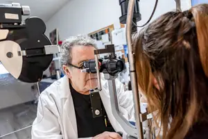 An elderly man wearing glasses and a lab coat examines a woman's eyes with a slit lamp in a clinical setting
