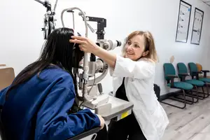 A smiling female optometrist is helping a female patient use an eye examination machine in a clinic.