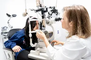 An adult woman wearing a white coat is examining the eyes of a young girl using an ophthalmoscope in a white room.