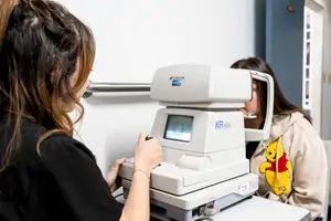 A woman uses a Topcon eye examination machine on another woman in an examination room.