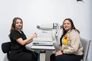 Two women are sitting in front of a white wall, one woman is operating an eye examination machine with the words 'KR 800' on it, and the other woman is smiling and wearing a Winnie the Pooh hoodie.