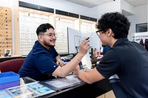 Two men in an optical shop, one with glasses pointing at a monitor while the other holds a pen, both smiling and focused on the task at hand.