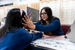 A woman in glasses is adjusting a girl's head in a shop with shelves and frames in the background.