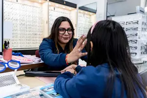 A woman is seen inside a store helping a customer with her glasses in front of a table with various items.