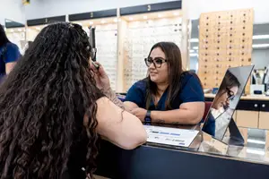 A woman with curly hair is adjusting her glasses while another woman is looking at her in a mirror, with an optical shop in the background.