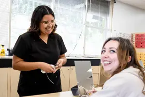 A woman in a black shirt is smiling and fixing the eyeglasses of a woman sitting in front of her in a classroom with a mirror and cabinets in the background.