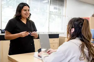 A woman wearing glasses is talking to another woman with a ponytail, who is sitting in front of a desk with a laptop and a mirror, in an office with glass windows and blinds.