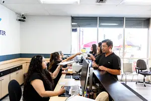 Inside an office, three women are seated in front of computers, while a man stands behind them, and another woman holds a paper. There is a surveillance camera in the corner and a window on the right side.