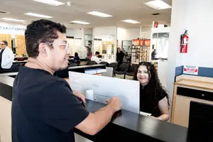 A man and a woman are smiling at a front desk in an office with a computer and a fire extinguisher on the wall