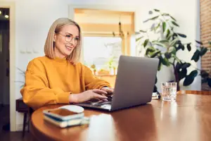 A woman wearing glasses sitting at a table using a laptop while smiling