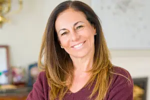 A woman with blonde hair and a maroon top is smiling and looking at the camera in a room with a fireplace