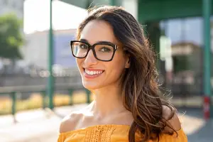 A woman with brown hair and glasses smiling at the camera, standing on a concrete pathway with a metal fence behind her