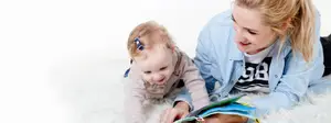 A woman and a baby are lying on a white fur carpet, the woman is holding a book and the baby is looking at the book with a smiling face