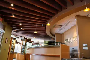 An indoor area resembling a reception area with a reception counter, a glass railing, and a wooden ceiling. The reception counter features a sign that reads 'McCorme Opticians.' The area is adorned with hanging lamps, a surveillance camera, and a glass wall. A glass cabinet is positioned near the wall, and a power outlet is attached to the wall.