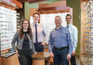 Four people are standing in a store with glasses on display, smiling at the camera.
