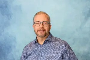 A man with glasses and a beard smiling and standing in front of a blue background