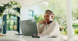 A woman sitting at a table with a laptop, looking at the camera with a smile, and a vase of flowers behind her.