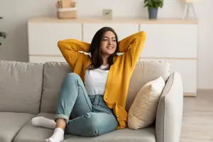 A woman sits on a sofa and smiles at the camera, wearing a yellow cardigan and white top, with a white pillow and couch behind her, and a wooden cabinet with a potted plant on top in the background.