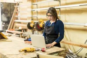 A woman wearing glasses and an apron is working on a laptop in a workshop.