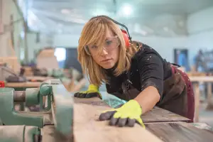 A woman in a workshop wearing safety gear working on a piece of wood