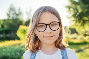 A young girl with glasses is smiling in a garden on a sunny day