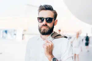 A man in a white shirt with a beard and sunglasses is adjusting his jacket while looking to the side