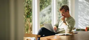 A man is sitting on a chair in front of a table with a laptop, a cup, a book, and a pen.