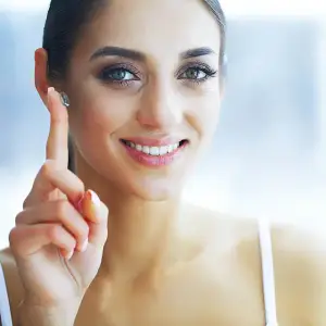 A smiling woman wearing a silver hearing aid in her ear, pointing at it with her finger