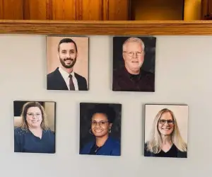 Five people are posing for a photograph on a white wall, with two men and three women, all wearing glasses and smiling, and the two men in suits.