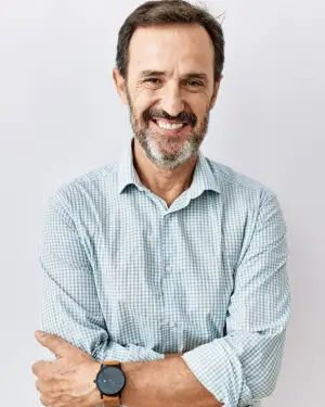 A smiling middle-aged man with a beard wearing a blue checkered shirt and a watch is posing for a photo.