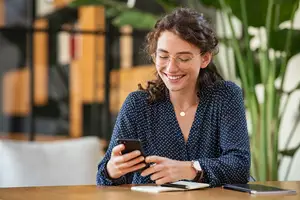 A woman with curly hair sits at a table, smiling while looking at her phone.