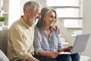 An older couple sitting on a couch, smiling, and looking at a laptop