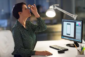 A woman is sitting in front of a desk in an office and is using eye drops
